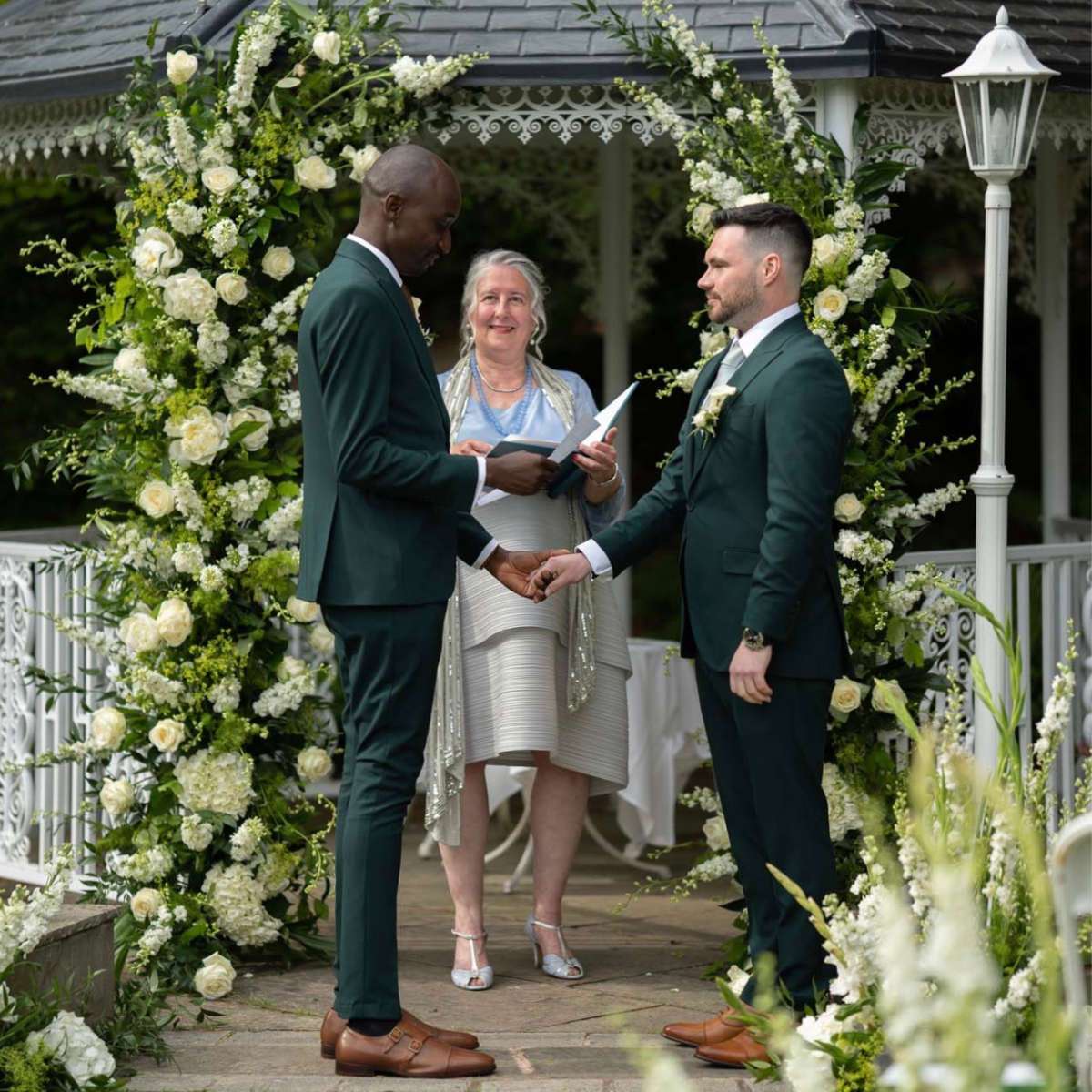 Catherine is standing under an arch of flowers during the ceremony of Emmett and Ayo who are both wearing dark green fitted suits and tan shoes. The couple are holding hands as Ayo is saying his vows.